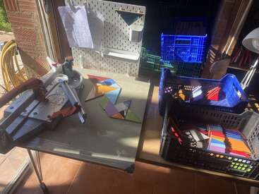 This image shows a workspace with a tile cutter, colorful triangular tiles, organized crates, a pegboard with papers, and sunlight streaming in from an open door.