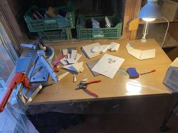 A well-lit workbench with tile cutting tools, scattered triangular tile pieces, pliers, gloves, instruction paper, crates of tiles, ruler, and other crafting materials.