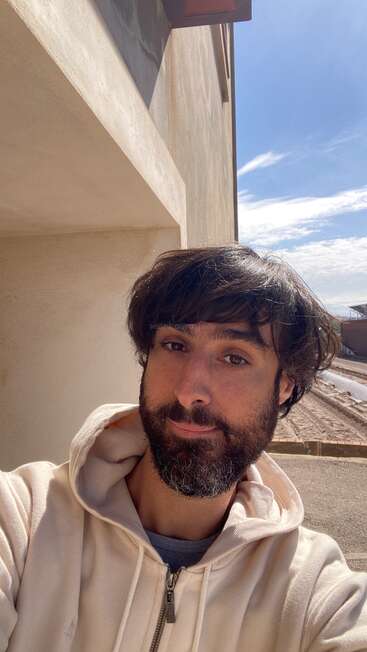 A bearded man in a cream hoodie takes a selfie outside next to a building. The sky is bright blue with some clouds, and sunlight shines.
