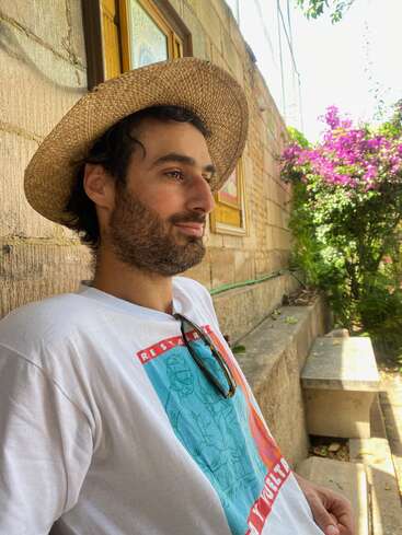 Un hombre con un sombrero de paja está recostado contra un muro de piedra; lleva una camiseta estampada y gafas de sol, y a su alrededor hay flores de un morado intenso y vegetación, mientras disfruta de un día soleado.