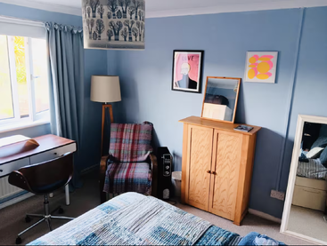 A cozy blue bedroom features a wooden desk, swivel chair, plaid-covered armchair, standing lamp, cabinet with mirror, artwork, window, and full-length mirror.