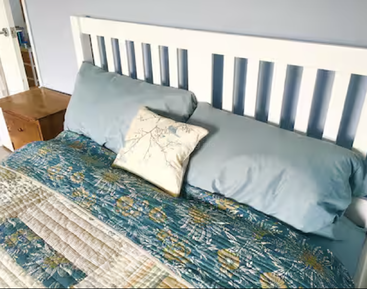 A neatly made bed with a white slatted headboard, blue pillows, a decorative floral cushion, and a colorful floral quilt. A wooden nightstand nearby.