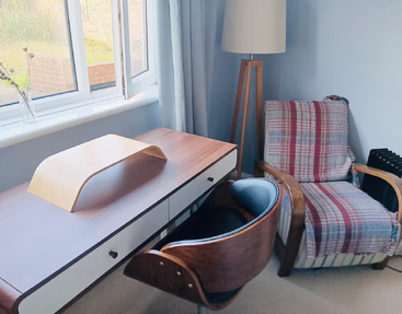 A cozy study nook with a wooden desk, stylish chair, armchair draped in a plaid blanket, standing lamp, and large window letting in sunlight.