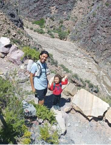 Un hombre y un niño, ambos con mochilas, sonríen mientras caminan por un cañón rocoso. El niño saluda alegremente, rodeado de escarpados acantilados y vegetación.