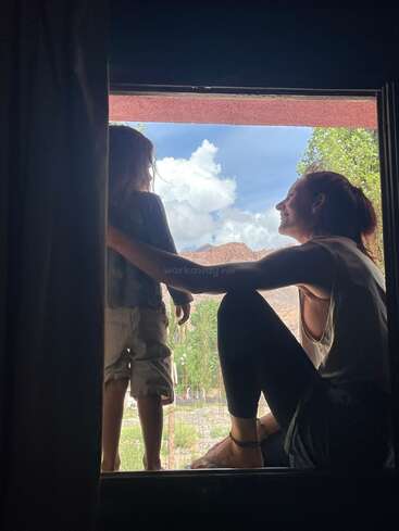 Una mujer y un niño sentados en el marco de una ventana, mirando al exterior, a un cielo azul brillante con nubes y montañas lejanas, compartiendo un momento de paz.