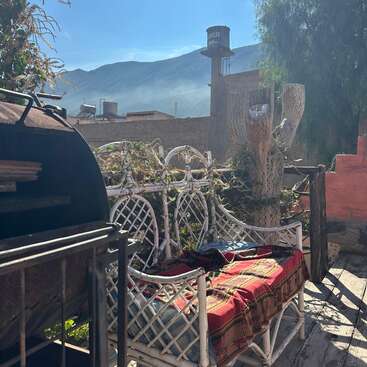 Patio rústico iluminado por el sol con banco de mimbre blanco, manta de colores, barbacoa, terraza de madera, plantas desérticas, montañas, torre de agua y un ambiente cálido y relajado por la mañana.