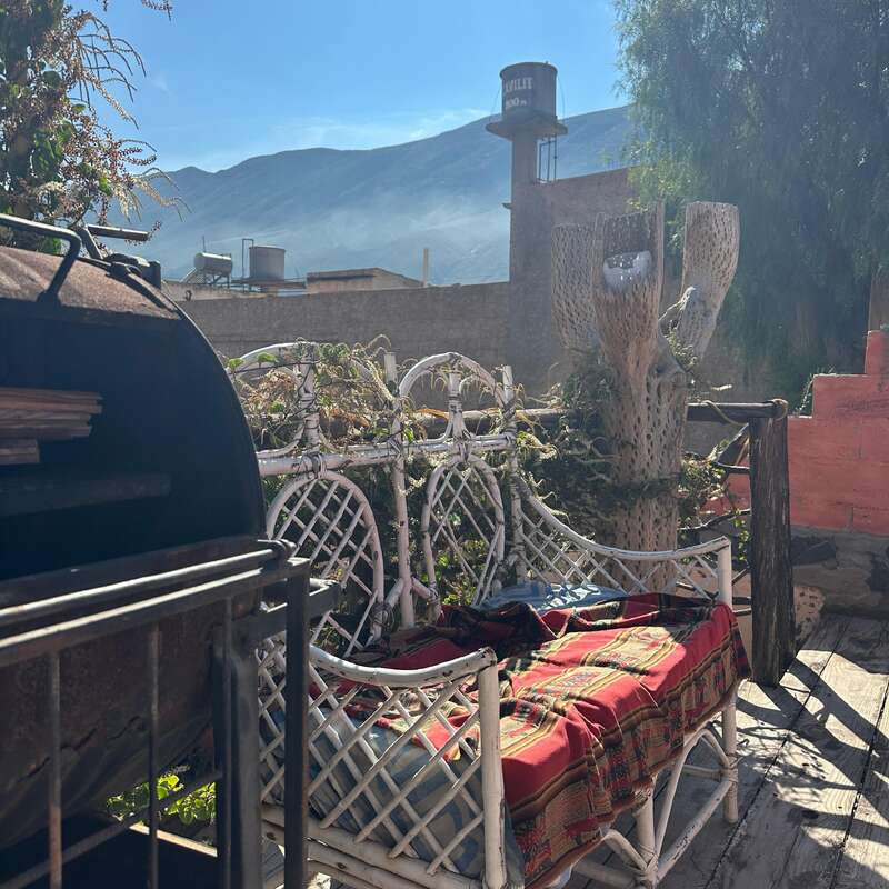 Patio rústico iluminado por el sol con banco de mimbre blanco, manta de colores, barbacoa, terraza de madera, plantas desérticas, montañas, torre de agua y un ambiente cálido y relajado por la mañana.
