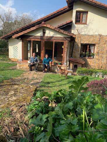 Das Bild zeigt eine ruhige Szene mit einem Paar und seinem Hund, die auf der Veranda eines charmanten Hauses sitzen, umgeben von üppigem Grün und einem klaren blauen Himmel.