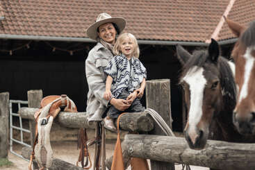 Uma mulher sorridente, usando um chapéu de cowboy, segura uma criança alegre em cima de uma cerca de madeira. Dois cavalos estão parados ali perto, com um estábulo e selas ao fundo.
