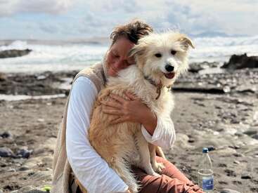 A woman hugs a fluffy, happy dog on a rocky beach. The ocean waves crash in the background. Both appear peaceful and content, enjoying the moment.