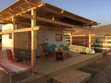 A cozy caravan sits under a wooden pergola, featuring two chairs, a table, a striped lounge chair, surfboards, and surrounded by a rustic, sunny landscape.
