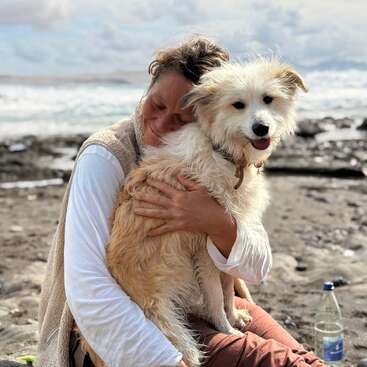 A woman hugs a fluffy, happy dog on a rocky beach. The ocean waves crash in the background. Both appear peaceful and content, enjoying the moment.