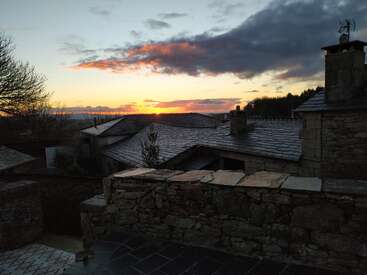 The image depicts a serene sunset scene featuring stone buildings with slate roofs, set against a backdrop of trees and a cloudy sky.