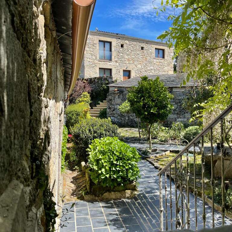 The image depicts a serene stone house with a lush garden, featuring a paved walkway, trees, and bushes, set against a bright blue sky with wispy clouds.