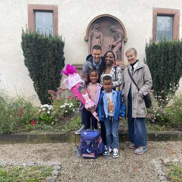 Eine glückliche fünfköpfige Familie steht draußen vor einem Gebäude und feiert einen besonderen Anlass mit Blumen, einem großen Schulkegel und einem Kinderrucksack.