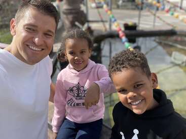 A man and two children smile happily for a selfie outdoors. They are near a pond with colorful decorations in the background, enjoying a sunny day together.
