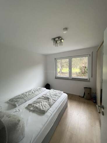 This image shows a small, minimalistic bedroom with light wooden floors, a double bed, white bedding, a window with a garden view, and simple decor.