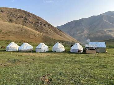 Five traditional yurts and a small metal structure sit on a green grassy plain, surrounded by mountains under a clear sky, horses grazing nearby.