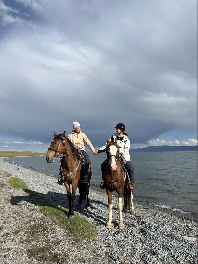 Two people on horseback ride along a pebbly lakeshore. They hold hands, smiling. Overhead, dramatic clouds stretch across the sky, creating a peaceful, scenic atmosphere.