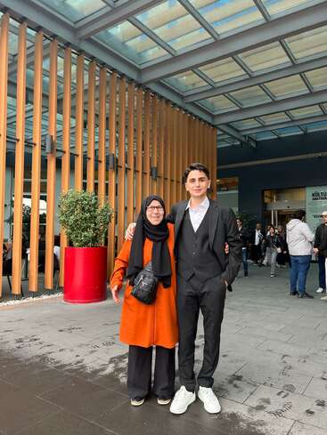 A smiling woman in an orange coat and headscarf stands next to a young man in a suit. They are outdoors, posing under a modern glass canopy.