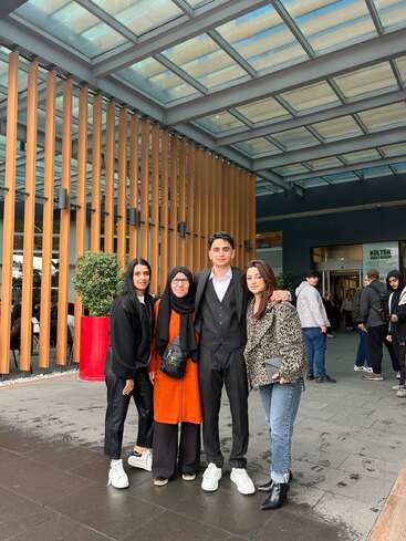 Four people posing together under a modern glass roof, smiling. They are stylishly dressed, standing close outside a building. Other people are seen in the background.
