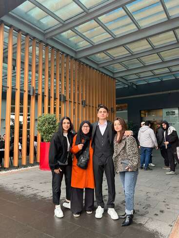 Four people pose closely together outside a modern building with wooden slats and a glass roof. They look happy, stylishly dressed, and seem to be enjoying the day.