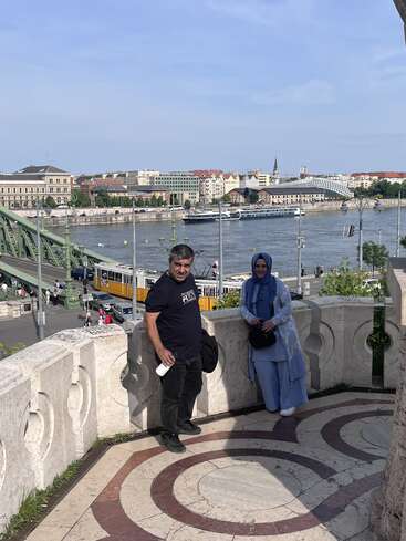 A man and woman stand on a stone terrace overlooking a river, tram, and cityscape with historic buildings and bridge on a sunny day.