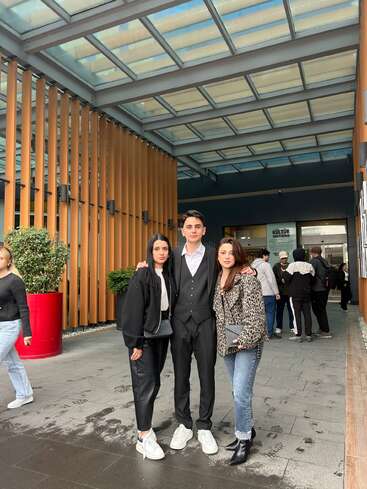 Three people pose together at the center of a modern, covered outdoor area with wooden panels, surrounded by others casually walking or standing in the background.