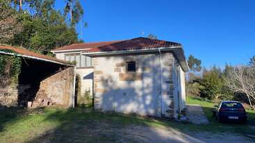 Uma casa rústica com telhado de telhas fica ao lado de um galpão de pedra. Um carro está estacionado nas proximidades. A cena é ensolarada, cercada por árvores e vegetação.