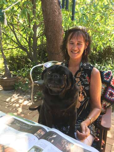 The image depicts a woman sitting in a chair with a black pug on her lap, surrounded by lush greenery, reading a book with a serene outdoor setting.