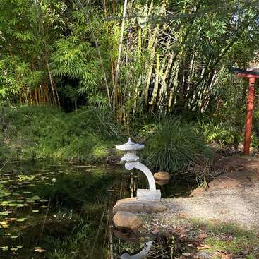 The image depicts a serene Japanese-style garden, featuring a tranquil pond, a white stone lantern, and lush greenery, with a red torii gate in the background.