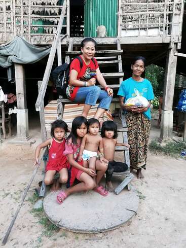 Un groupe de cinq enfants et deux femmes posent devant une maison rustique sur pilotis. Le cadre semble rural et tout le monde a l'air détendu et heureux.