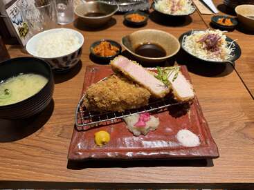 Un repas japonais composé de côtelettes de porc panées (katsu), de chou râpé, de riz, de soupe au miso, de légumes marinés, d'une sauce pour tremper, de moutarde et d'assaisonnements, le tout joliment présenté sur une table en bois.