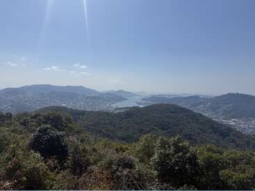 Un paysage pittoresque composé de collines verdoyantes, d'une ville au loin et d'un plan d'eau paisible, sous un ciel bleu limpide baigné par les rayons du soleil.
