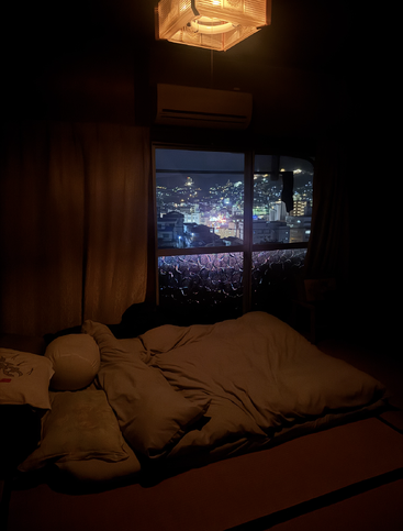 A cozy, dimly lit bedroom with a futon bed, soft pillows, and a cityscape view through the window, glowing with colorful night lights outside.