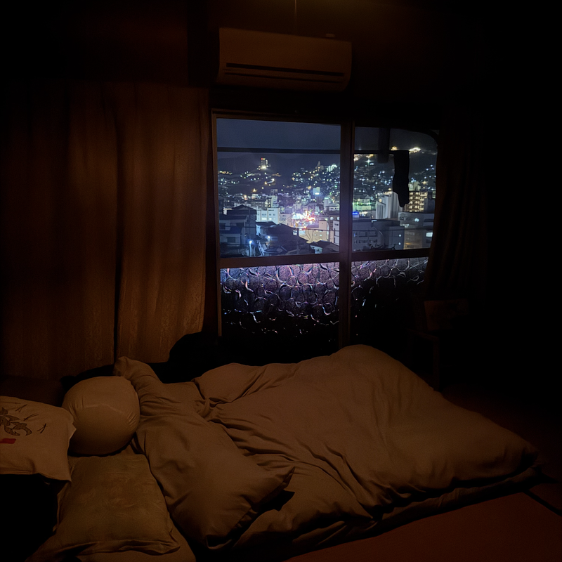 Une chambre intime et tamisée, avec un lit futon, des oreillers moelleux et une vue sur la ville depuis la fenêtre, illuminée par les lumières colorées de la nuit.