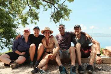 The image depicts a group of five individuals, likely family members, sitting together on a dirt surface, posing for a photo with their arms around each other.