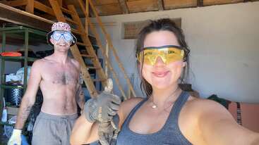 The image depicts a man and woman in a garage, both wearing safety goggles and gloves, with the woman giving a thumbs-up. The man is shirtless, while the woman wears a tank top.
