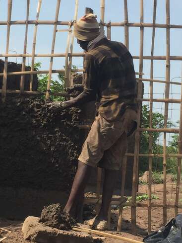 A imagem mostra um homem construindo uma parede usando lama, em pé sobre uma pilha de lama com uma estrutura de bambu ao fundo em um dia ensolarado.
