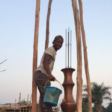 A imagem mostra um homem em pé na frente de uma estrutura alta, segurando um balde azul, com um grande pote de barro por perto, em um cenário de céu sereno.
