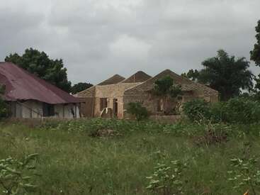 A imagem mostra um campo gramado com uma casa em construção ao fundo, com telhado e paredes parcialmente concluídos, cercada por árvores e um céu nublado.