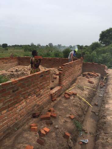 A imagem mostra dois indivíduos construindo uma parede de tijolos em uma fundação de terra, com uma pessoa segurando um tijolo e a outra trabalhando na estrutura da parede.