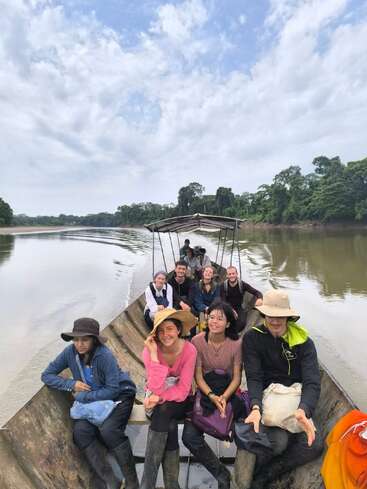 A group of people sit in a wooden boat, traveling down a wide, calm river surrounded by lush green forest, under a cloudy blue sky.