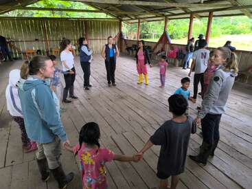 A group of adults and children stand in a wooden shelter, holding hands in a large circle, engaged in a group activity or game, surrounded by nature.
