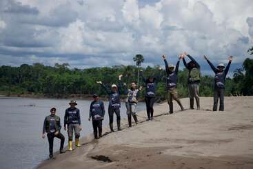 Eight people stand on a sandy riverbank, surrounded by lush green forest. Some have arms raised in excitement. The sky is cloudy, suggesting a warm day.