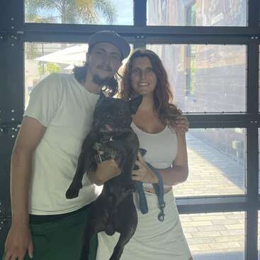 Um homem e uma mulher estão juntos, sorrindo, segurando um buldogue francês preto. Eles estão dentro de casa, perto de uma janela ensolarada, com uma palmeira visível do lado de fora.