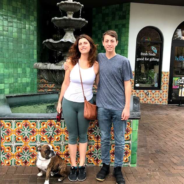The image depicts a woman, a boy, and a dog standing in front of a fountain and storefront, with the woman\'s arm around the boy\'s shoulder.