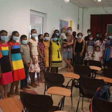 Das Bild zeigt eine bunte Gruppe von Menschen, darunter Kinder und Erwachsene, die in einem Klassenzimmer stehen, alle mit Gesichtsmasken, vor denen Tische und Stühle aufgestellt sind.