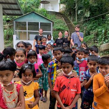 A group of children, some wearing masks, stand outdoors in colorful clothes. Behind them, a few adults and greenery are visible, suggesting a school or community setting.