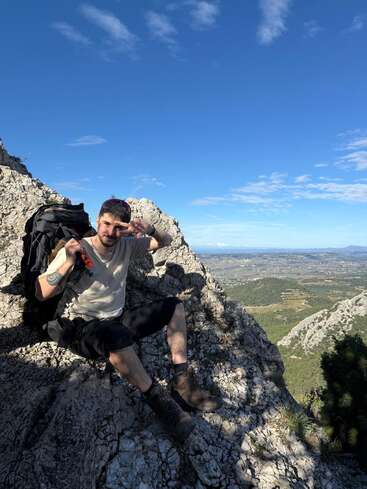 A man sits on a rocky mountain peak, wearing boots and outdoor gear, with a backpack beside him. He flashes a peace sign, enjoying the scenic view.